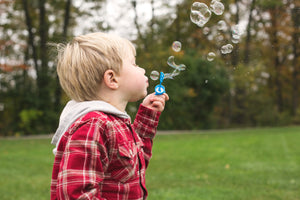 A young boy wearing a red shirt blows bubbles outside stood on green grass