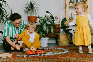 A mother and her two young children playing at home surrounded by green plants. The children are dressed in yellow clothes and the boy is playing with a train set